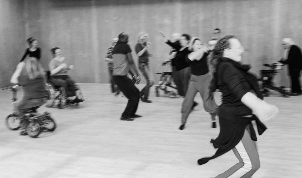 Wilde Tanz-Improvisation bei den Proben des Misch-MASH-Projektes "Spüren hinterlassen": Menschen mit und ohne körperliche Einschränkungen, teilweise mit Rollstuhl, Rollator und anderen Hilfsmitteln bewegen sich frei auf einer Tanzfläche.